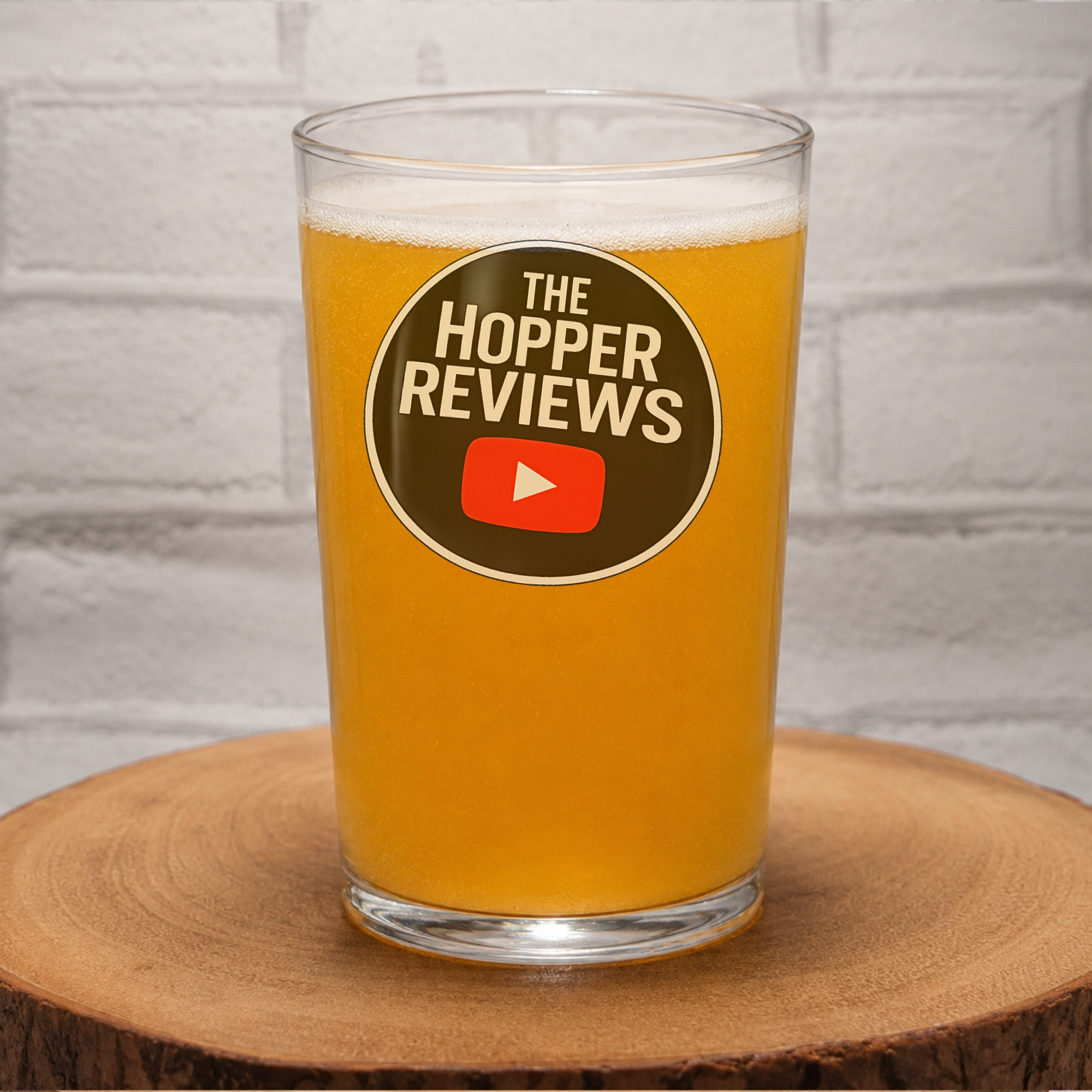 A clear pint glass filled with golden beer, standing on a round wooden slice in front of a white brick wall. The front of the glass has a circular brown logo reading 'THE HOPPER REVIEWS' with a red play button icon.