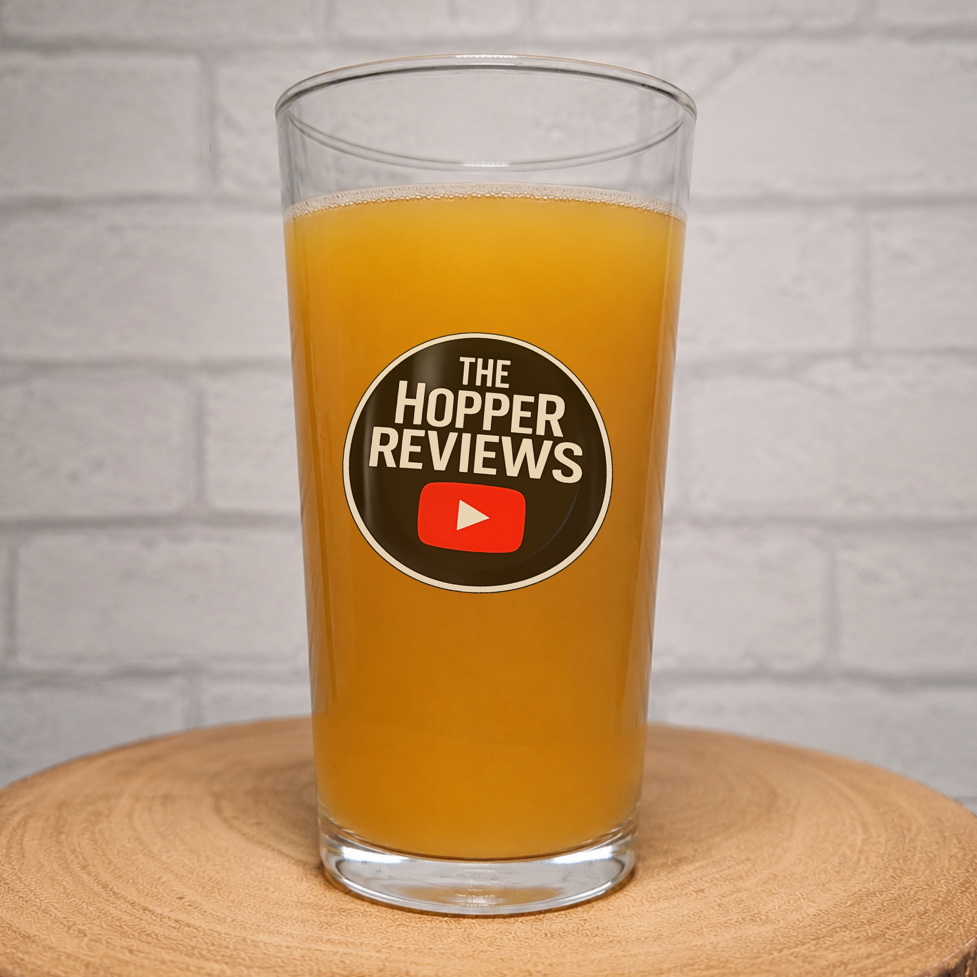 A clear pint glass filled with orange beer, featuring a circular black, white, and red 'The Hopper Reviews' logo on the front, set on a round wooden coaster against a white brick wall background.