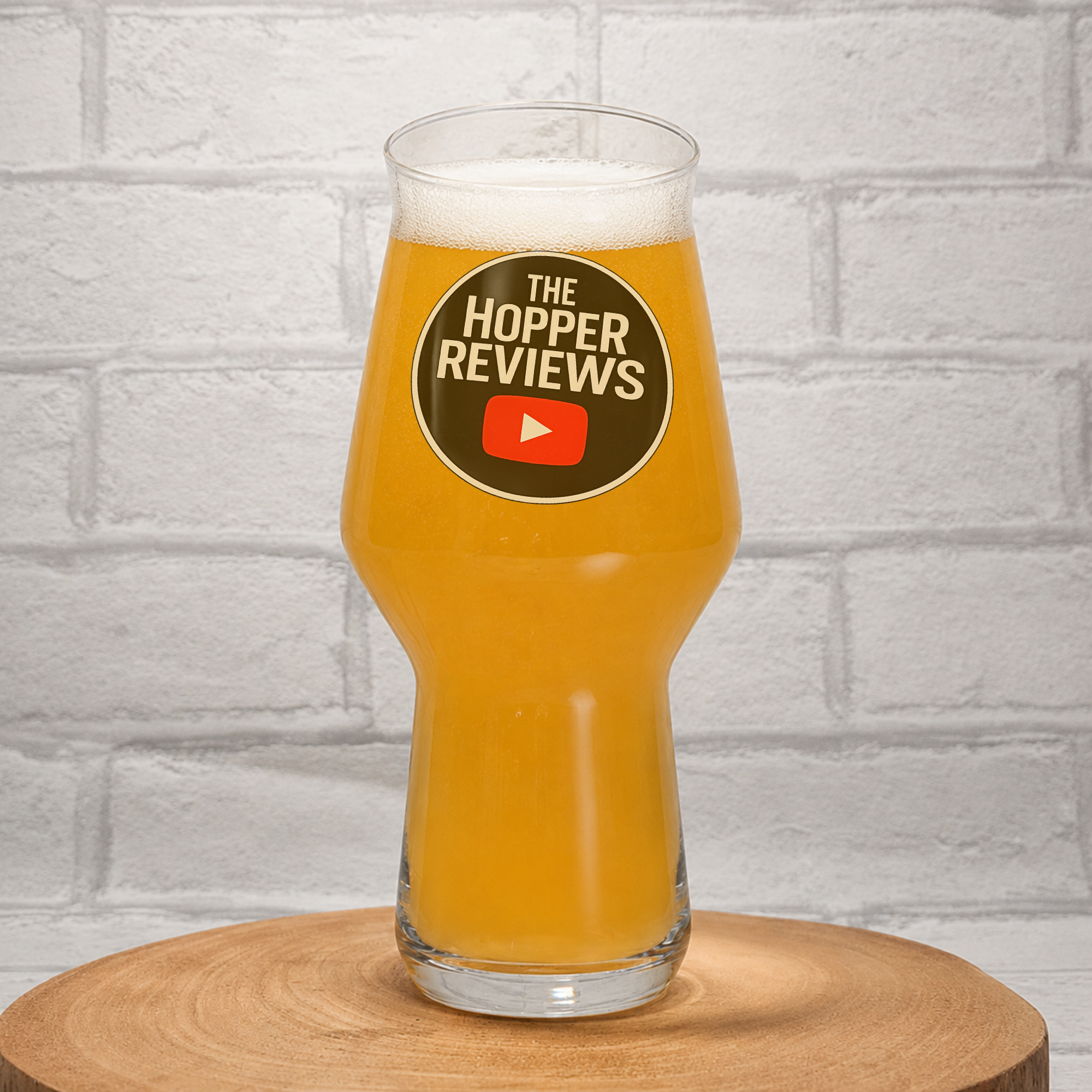 A clear pint glass filled with orange beer, featuring a circular black, white, and red 'The Hopper Reviews' logo on the front, set on a round wooden coaster against a white brick wall background.