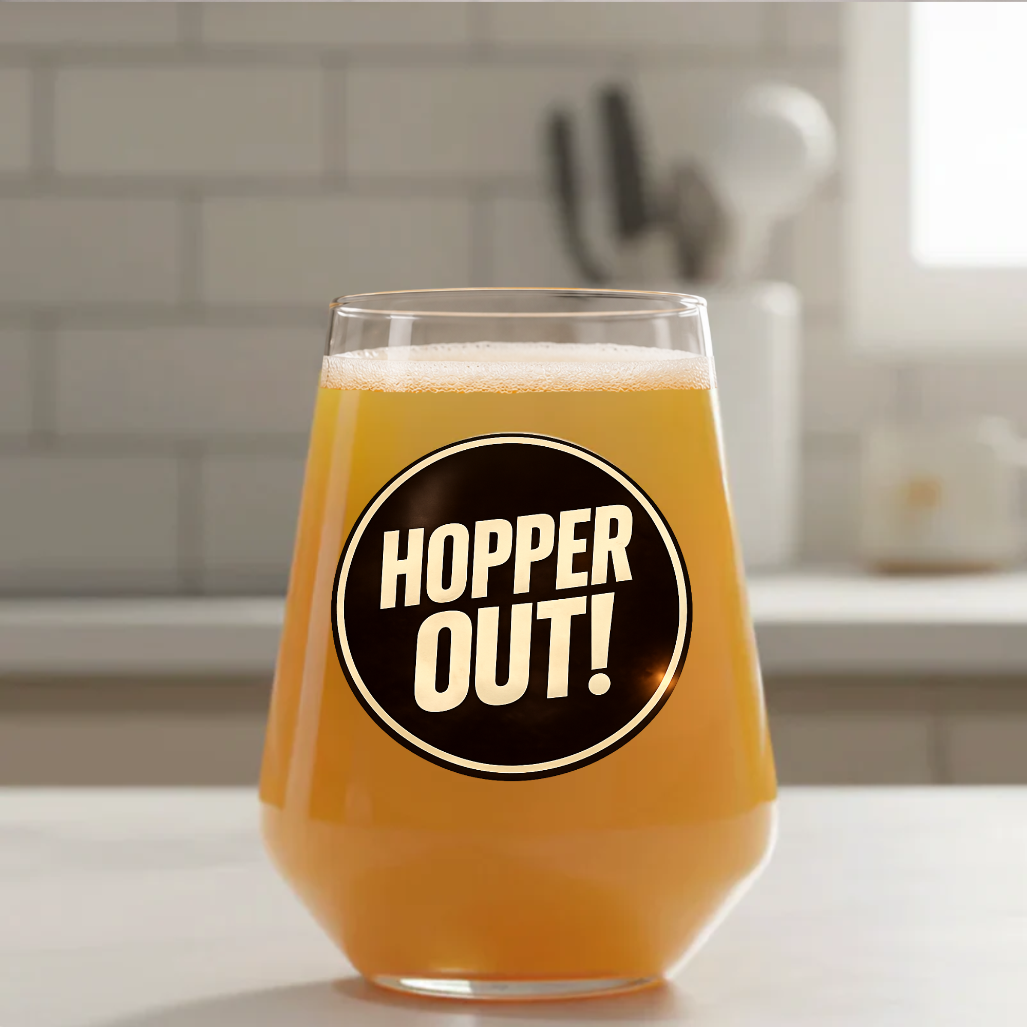 A clear stemless drinking glass filled with amber beer, placed on a countertop in a blurred kitchen setting. The front of the glass has a large black circular graphic with the words “HOPPER OUT!” in cream lettering.