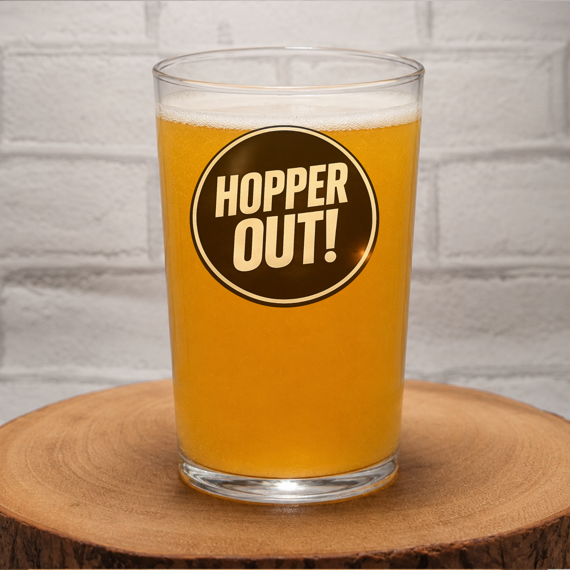 A clear pint glass filled with golden beer, featuring a circular brown label with the words 'HOPPER OUT!' in cream lettering, set on a wooden slice against a white brick wall background.