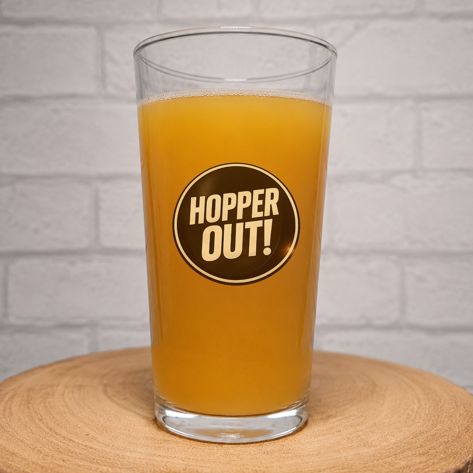 A clear pint glass filled with orange beer, featuring a circular black and gold 'HOPPER OUT!' logo on the front, set on a wooden coaster against a white brick wall background.