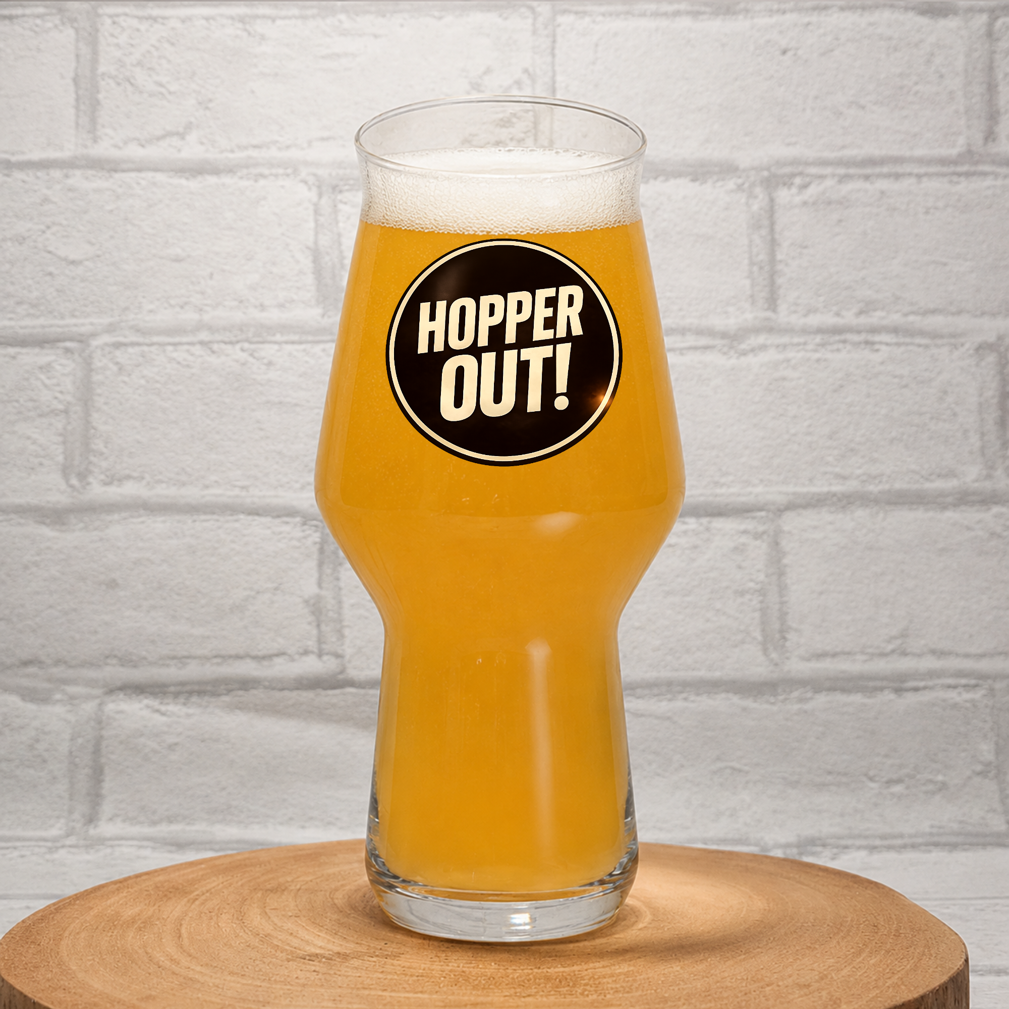 A clear pint glass filled with golden beer, standing on a round wooden coaster against a white brick wall. The glass features a black circular sticker with the text 'HOPPER OUT!' in white.